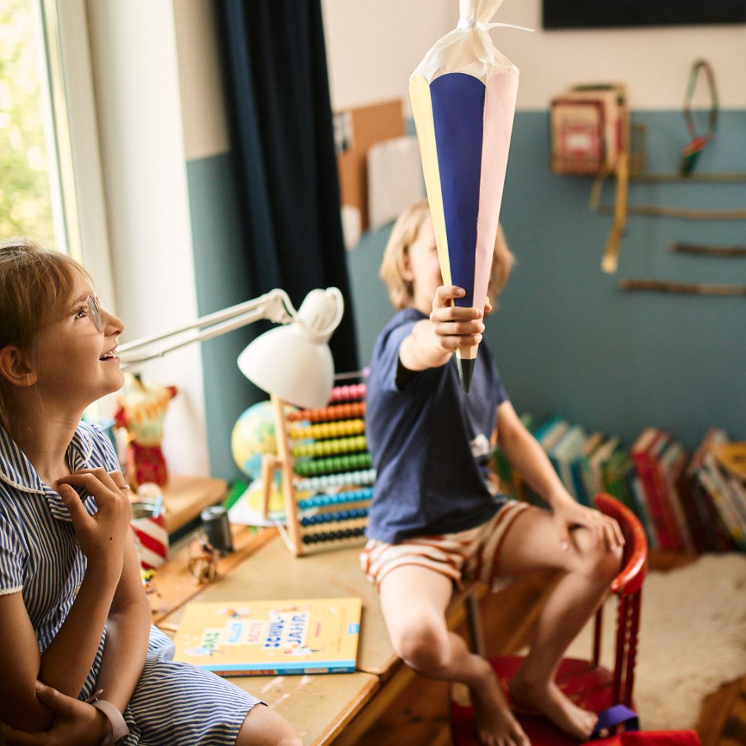 Zwei Kinder in einem farbenfrohen Zimmer genießen das Buch „Das allerbeste Schuljahr“ vom Zuckersüß Verlag. Das eine hält einen großen Papierstrauß in der Hand, während das andere lächelt, umgeben von Büchern und Spielsachen – eine fröhliche Szene, die von Schulzeiten inspiriert ist.