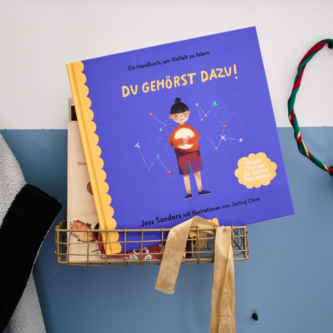 Das blau eingebundene Kinderbuch „Du gehörst dazu!“ vom Zuckersüß Verlag zeigt auf dem Einband ein Kind, das eine leuchtende Kugel in der Hand hält. Dieses farbenfrohe Bilderbuch, das Vielfalt feiert, liegt in einem Drahtkorb zusammen mit weiteren Büchern, einem Band sowie einer geflochtenen Kordel.