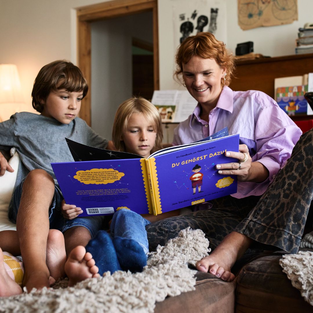 Eine Frau und zwei Kinder sitzen auf einem Sofa und lesen gemeinsam das Buch „Du gehörst dazu!“ vom Zuckersüß Verlag. Sie lächeln und sind ganz in die Lektüre vertieft, im Hintergrund ist eine gemütliche Wohnkulisse zu sehen.