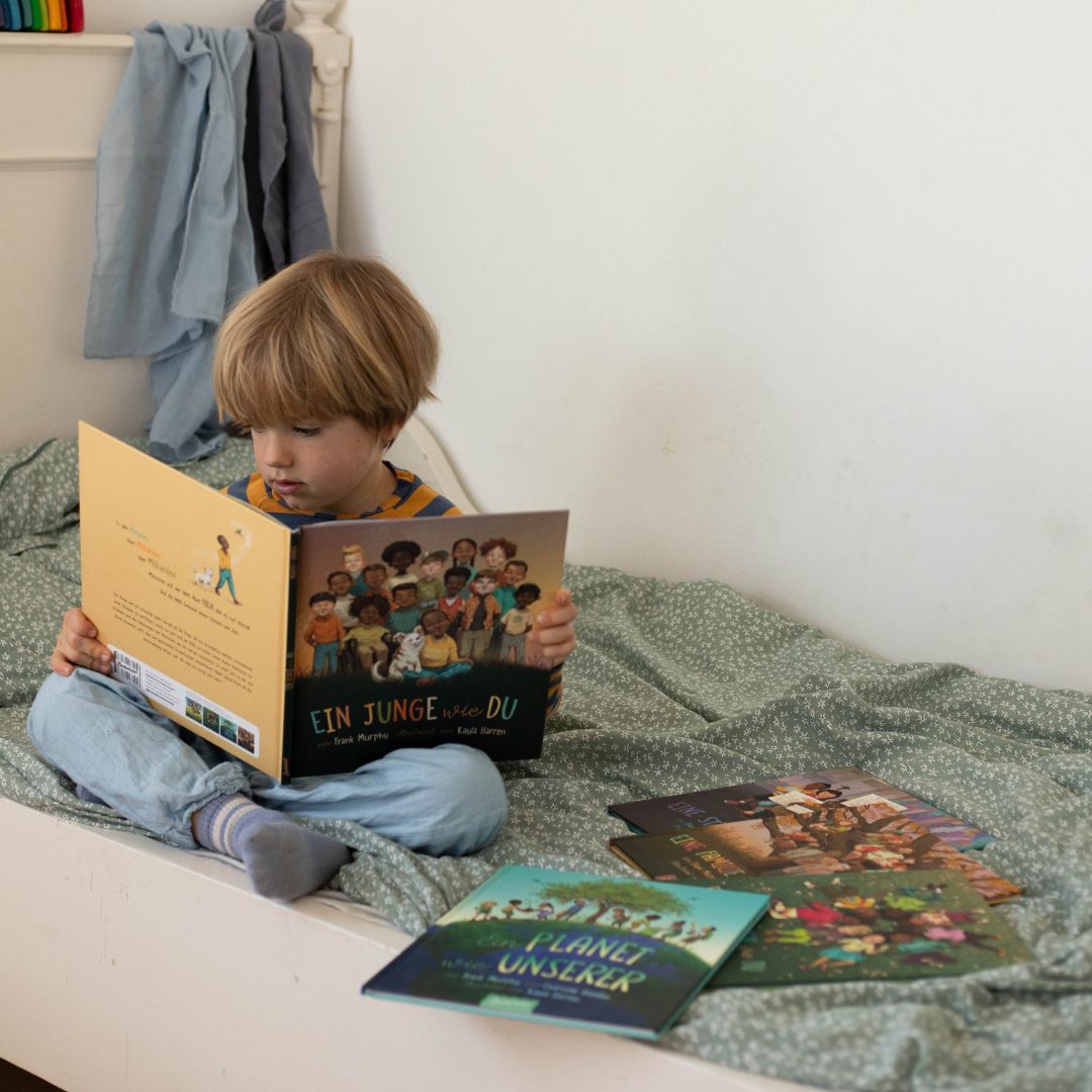 Ein kleines Kind sitzt im Schneidersitz auf einem Bett und liest ein Bilderbuch aus der Eine Welt wie diese - Jungs-Kollektion des Zuckersüß Verlags. Drei weitere bunte Bücher liegen neben ihnen. Das Zimmer ist einfach eingerichtet und hat eine grün gemusterte Decke.