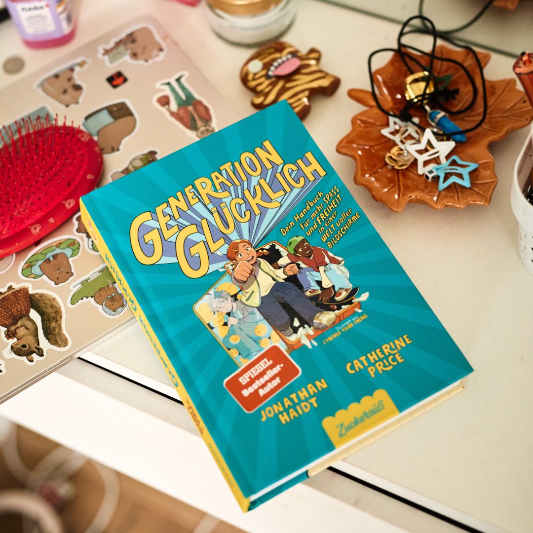 Das deutsche Buch "Generation Glücklich" aus dem Zuckersüß Verlag liegt auf einem unordentlichen weißen Schreibtisch, auf dem Aufkleber, eine rote Haarbürste, Schmuck und Schmuckschalen in Tierform liegen. Es beschäftigt sich mit Themen wie soziale Medien und Bildschirmzeit.