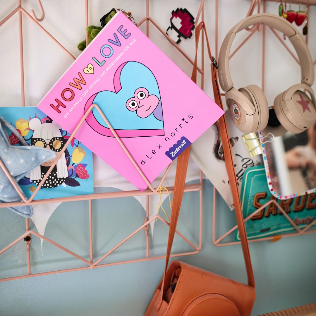 Ein rosa Drahtgitter hält ein rosa Buch mit dem Titel „How to love“ vom Zuckersüß Verlag, das mit einem Comic-Herz verziert ist und Inspiration für Dating und Liebe bietet, neben beigen Kopfhörern, einer braunen Handtasche, einer Postkarte, Stoffartikeln und einem Handy mit einer pastellfarbenen Hülle.