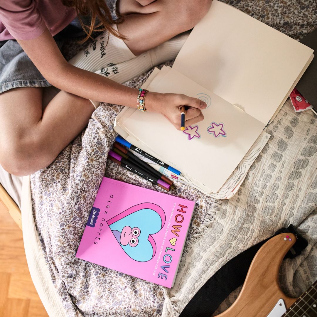 Eine Person sitzt auf einem Bett und zeichnet Sterne in ein Skizzenbuch, daneben liegen Filzstifte. Auf dem Bett liegen außerdem eine E-Gitarre und ein rosa Buch mit dem Titel „How to love“ vom Zuckersüß Verlag, das auf Beziehungen und Dating hindeutet. Die Kulisse wirkt ungezwungen und kreativ.