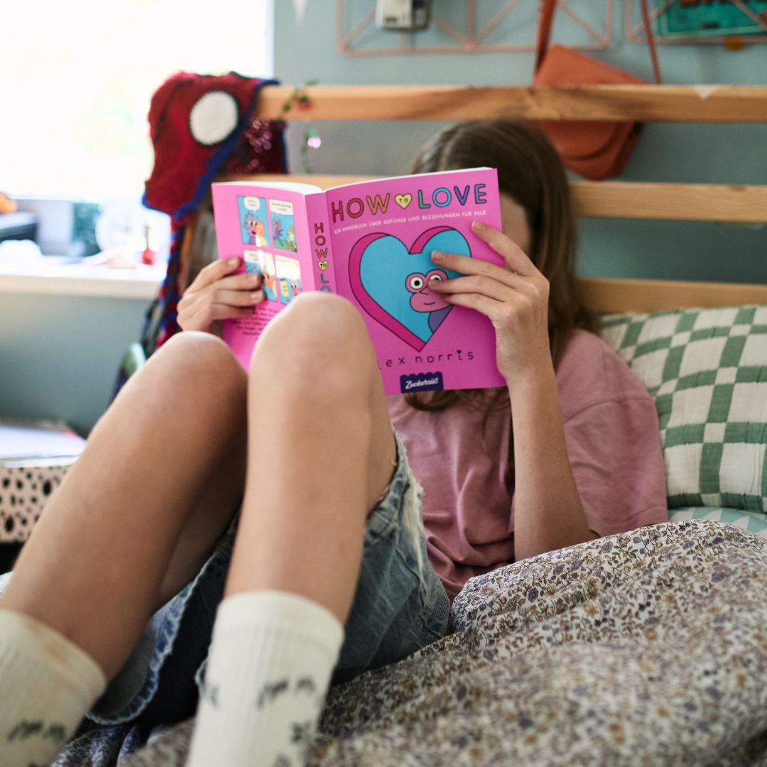 Eine Person sitzt auf einem Bett, hält das rosa Buch „How to love“ vom Zuckersüß Verlag in der Hand und liest darin; auf dem Einband ist ein gezeichnetes Herz abgebildet. Ihr Gesicht ist größtenteils hinter dem Buch verborgen, während sie sich in Ratschläge zu Dating und Beziehungen vertieft.