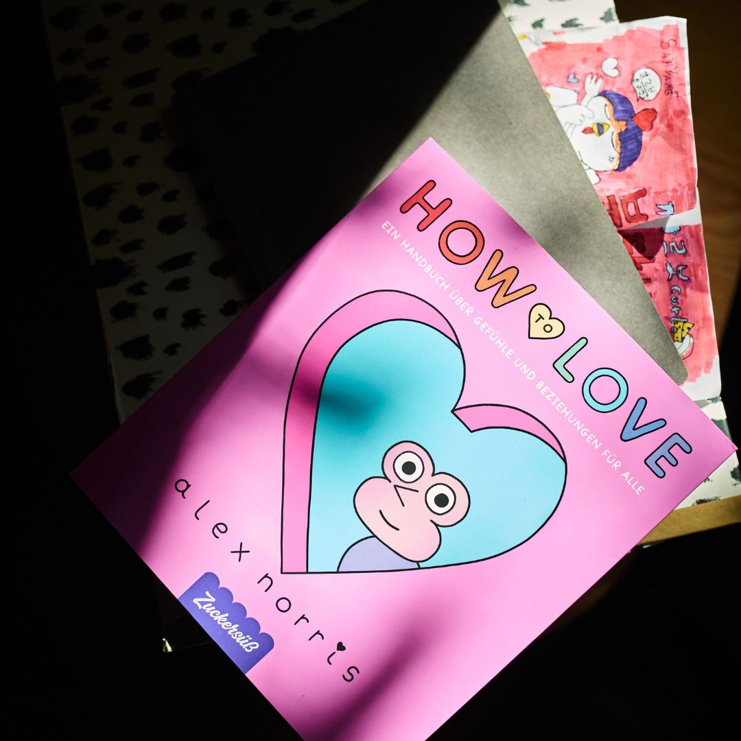 Ein rosa Buch mit dem Titel *How to love* vom Zuckersüß Verlag, dessen Einband mit einem blauen Herz und einem Cartoon-Gesicht verziert ist, liegt auf einem Tisch im Halbschatten – ein bezaubernder Ratgeber, ideal für alle, die sich mit Dating und moderner Liebe beschäftigen.