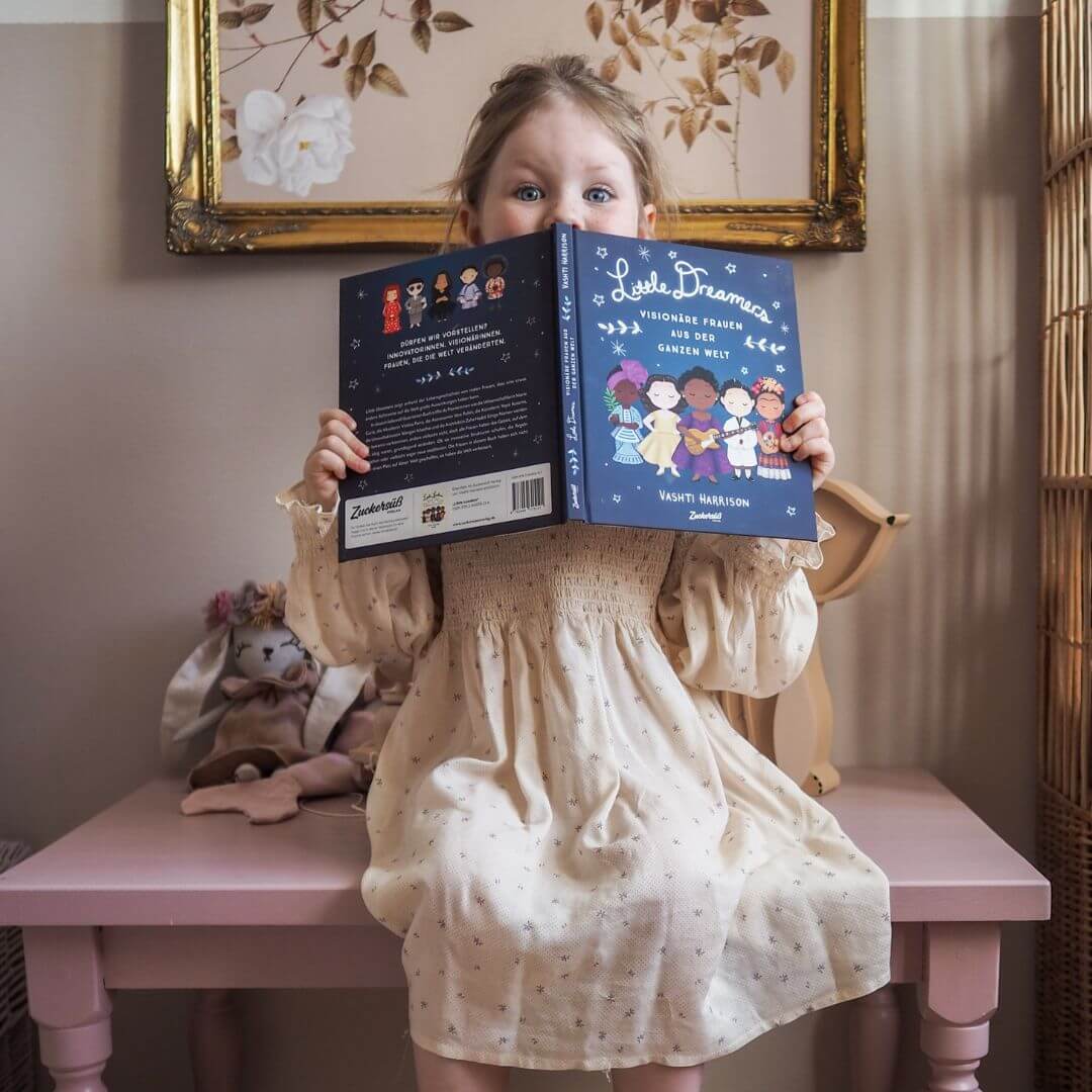 Ein junges Mädchen in einem cremefarbenen Kleid sitzt auf einem rosafarbenen Tisch und hält das Magic Dreamer Set aus dem Zuckersüß Verlag in der Hand - so, dass nur ihre Augen darüber schauen.