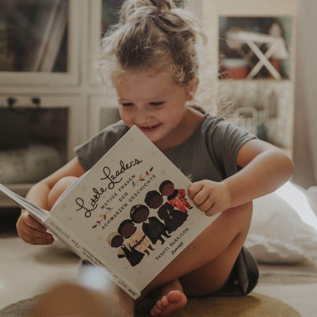 Ein junges Mädchen sitzt barfuß auf dem Boden und liest lächelnd das Magic Dreamer Set vom Zuckersüß Verlag - eines von vielen inspirierenden Büchern für Mädchen. Sonnenlicht strömt herein und schafft eine gemütliche Atmosphäre.