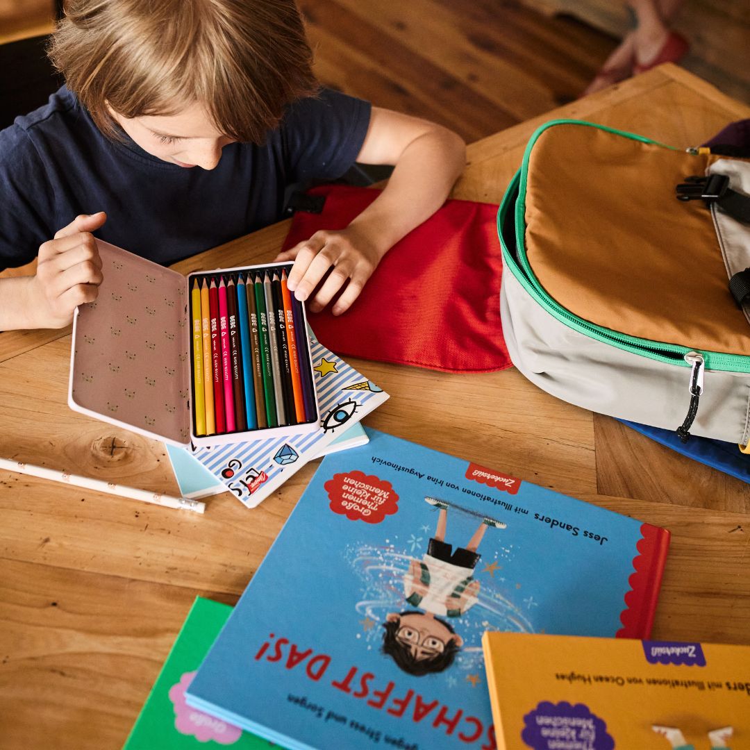 Ein Kind sitzt an einem Holztisch mit dem „Schulstarter-Paket“ des Zuckersüß Verlags – einer Dose mit Buntstiften, bunten Büchern, einem Federmäppchen und einem offenen Rucksack –, was die Vorfreude und Zuversicht beim Schulanfang einfängt.