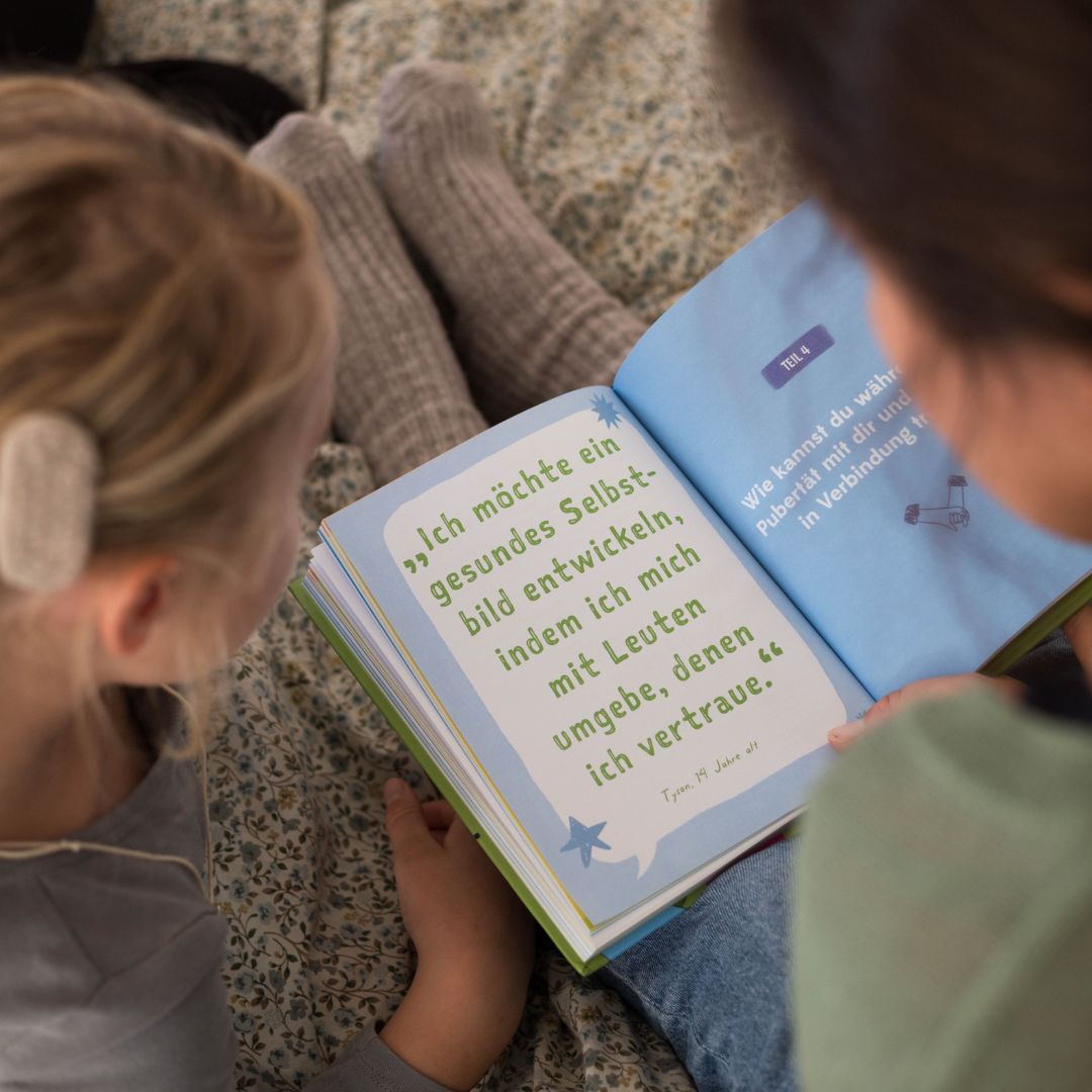 Zwei Kinder sitzen zusammen auf einem Bett und lesen "Von Null auf Pubertät" vom Zuckersüß Verlag. Ein Kind trägt ein Hörgerät. Die Seiten zeigen einen deutschen Text über Selbstverständnis und Vertrauen, beleuchtet von weichem, natürlichem Licht.